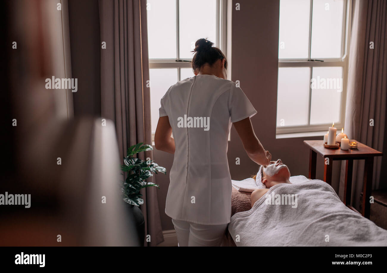 Beautician applying herbal face mask to female lying on massage table in beauty spa. Female performing a beauty treatment on woman client at health sp Stock Photo