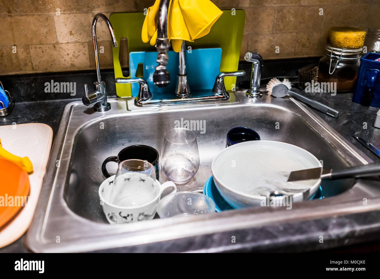 Closeup of dirty dishes in stainless steel sink kitchen by cleaning tools, cutting boards, brick wall Stock Photo