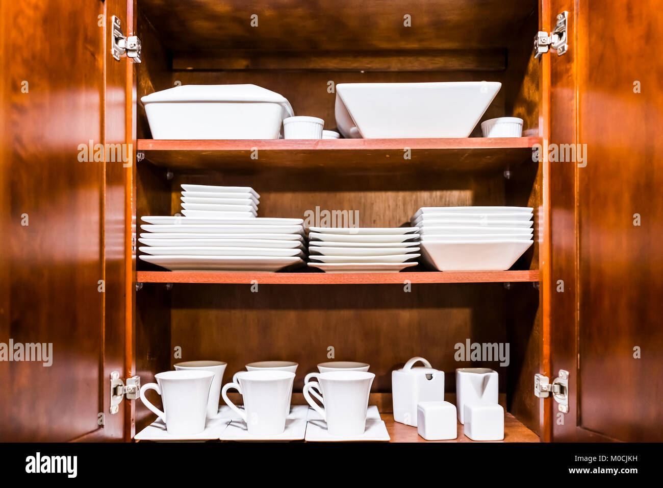 Open wooden kitchen cabinet door cupboard with many white dishes, plates, cups on shelves closeup Stock Photo