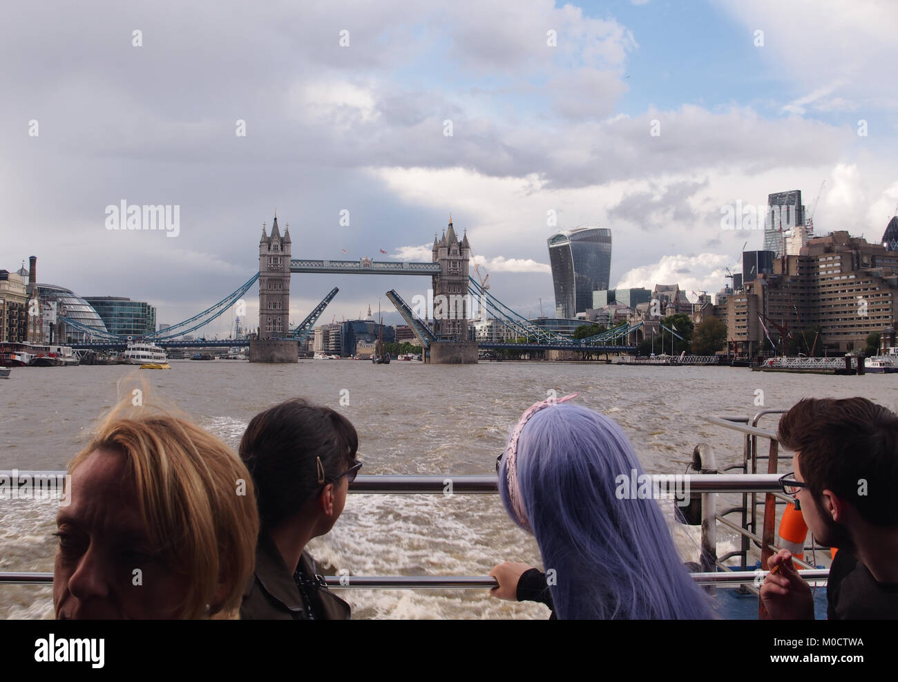 A view from the back of a Thames River passenger cruiser looking back to a raised Tower Bridge with four passengers in the foreground Stock Photo