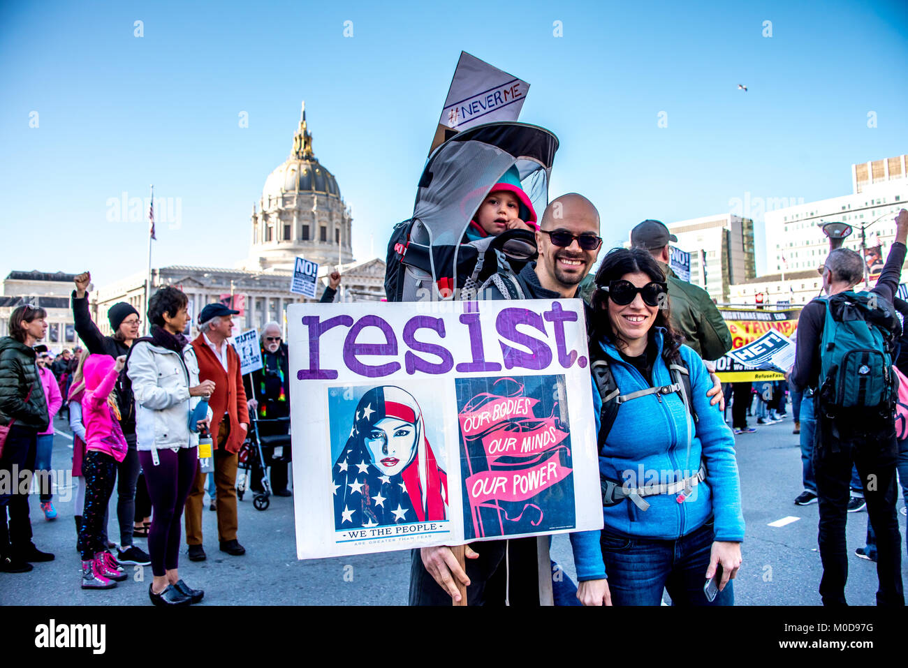 San Francisco, California, USA. 20th January, 2018. The 2018 Women's March in San Francisco, organized by Women's March Bay Area. A family prepares to march, mom and dad with baby on back in a child carrier backpack with its own sign reading #neverme and the father holds a sign that reads 'resist,' which includes the 'Our bodies, our minds, our power' and 'We the people' signs made popular during the previous women's march. Stock Photo