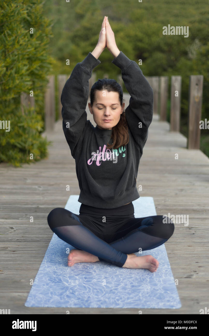 woman practicing yoga on a mat outside on a wooden boardwalk, crossed legged lotus pose. Stock Photo