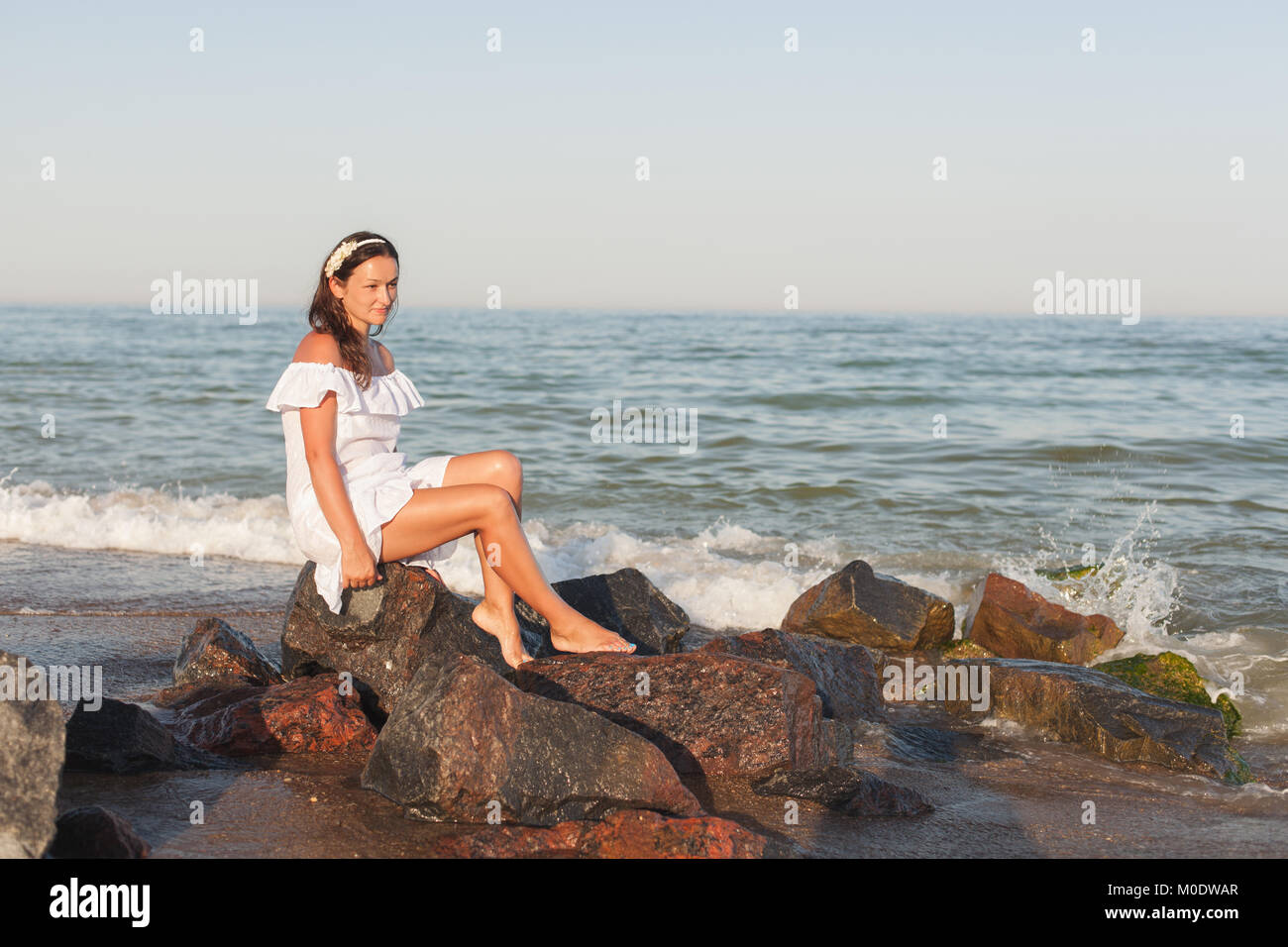 Young woman in white dress is sitting on stones on the seashore Stock ...