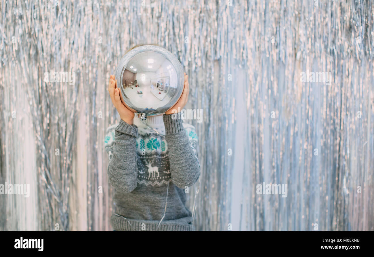 Boy standing in front of silver tinsel decorated wall holding a giant Christmas bauble Stock Photo