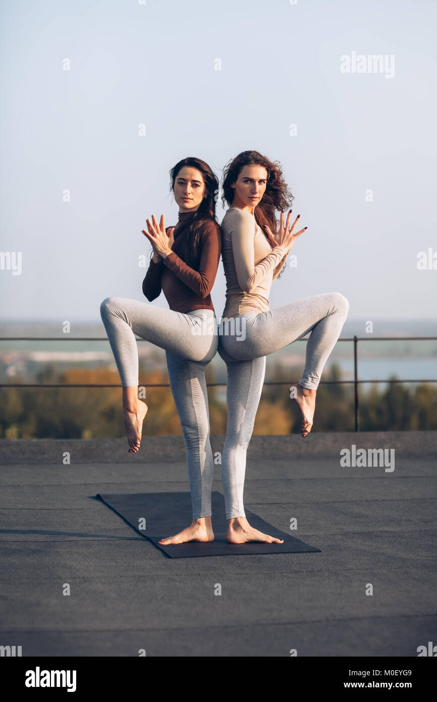 girls in standing couple yoga pose back to back on the roof outdoors M0EYG9