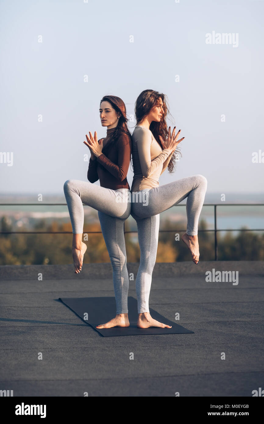 Girls in standing couple yoga pose back to back on the roof outdoors. Two beautiful women are concentrated on doing exercise while standing with raise Stock Photo