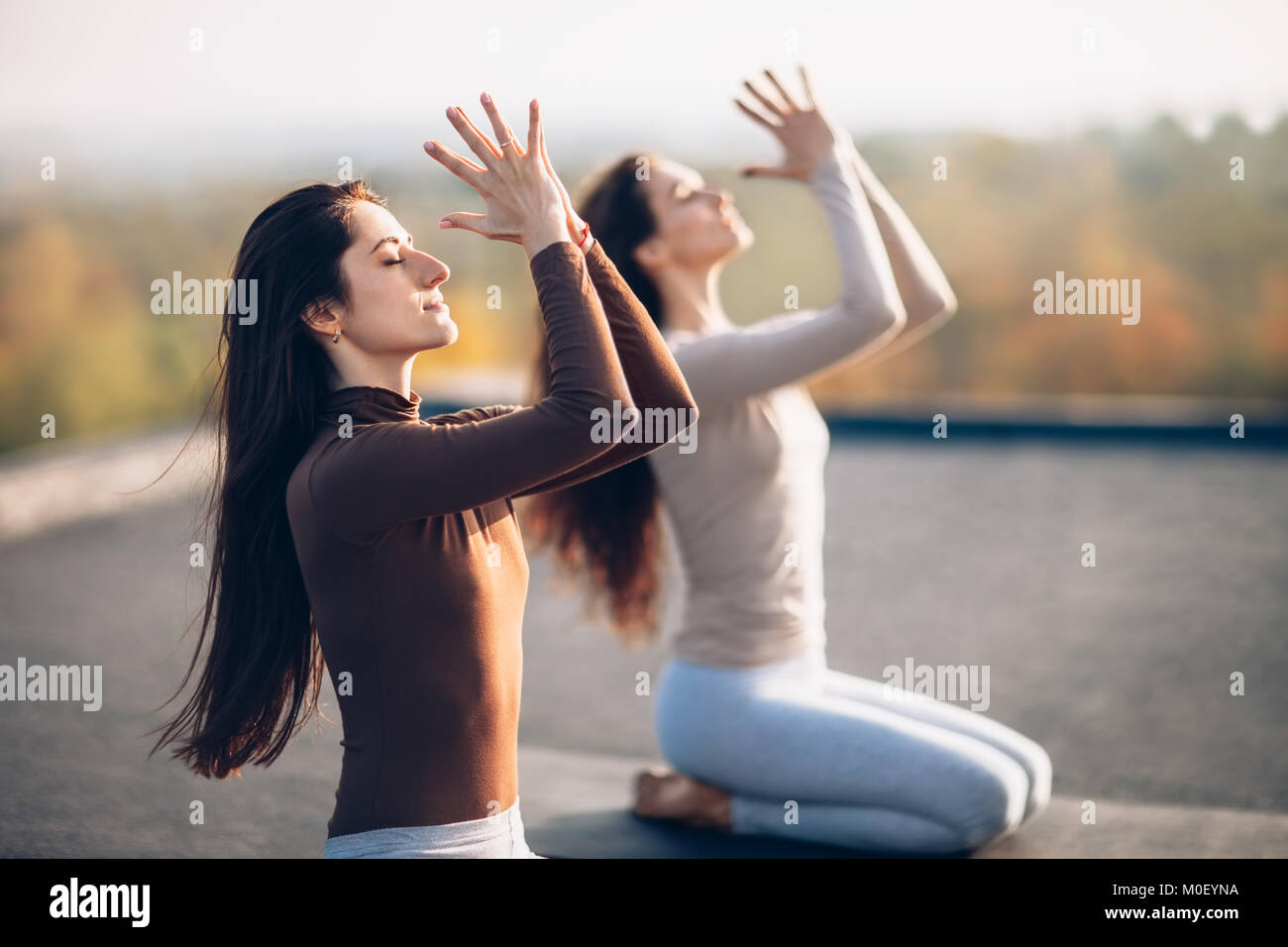 Two young beautiful women doing yoga asana, sitting in vajrasana exercise on the roof outdoor. Girlfriends perform a comfortable pose of eastern pract Stock Photo