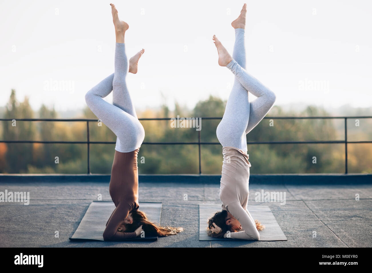 Two young women doing double yoga asana supported headstand on the roof outdoors. Salamba Sirsasana. Complex balanced posture for a healthy body and s Stock Photo