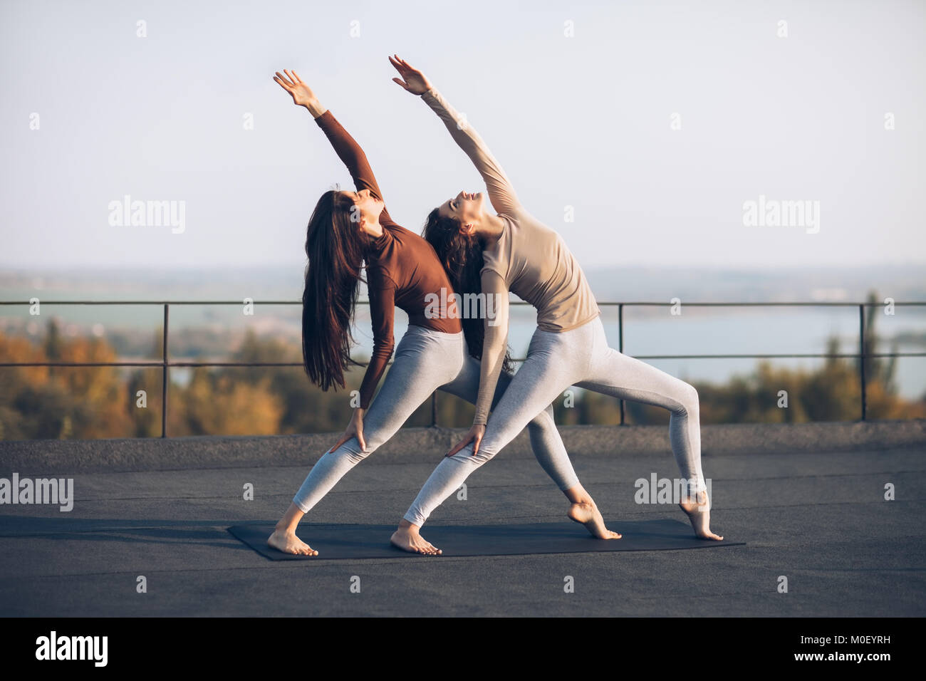 Girls synchronously perform double pose warrior with raised hand, on the roof outdoors. Beautiful yoga asana with elements of acrobatics. Healthy life Stock Photo