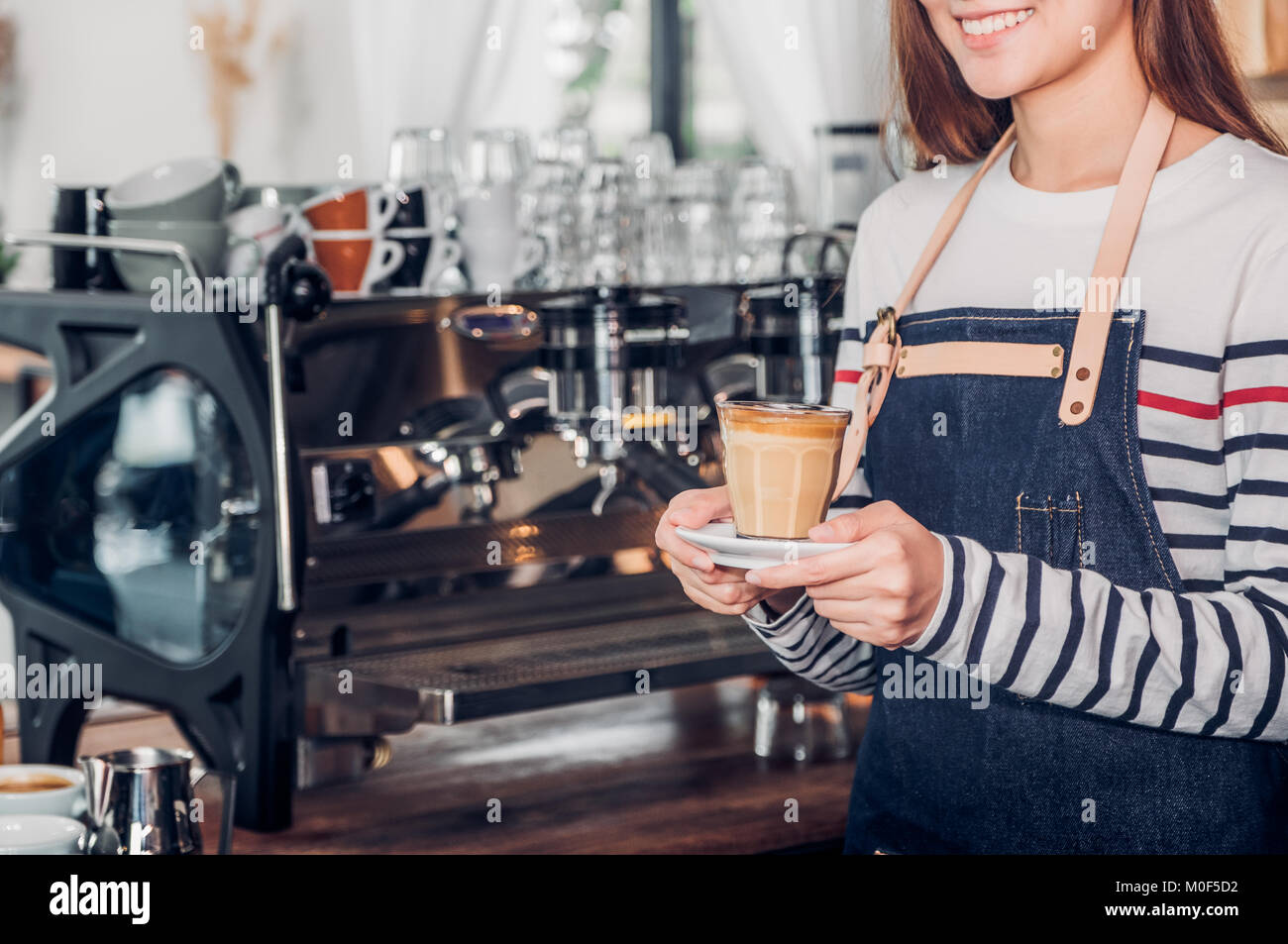 asia woman barista wear jean apron holding hot coffee cup served to customer with smiling face at bar counter,Cafe restaurant service concept.waitress Stock Photo