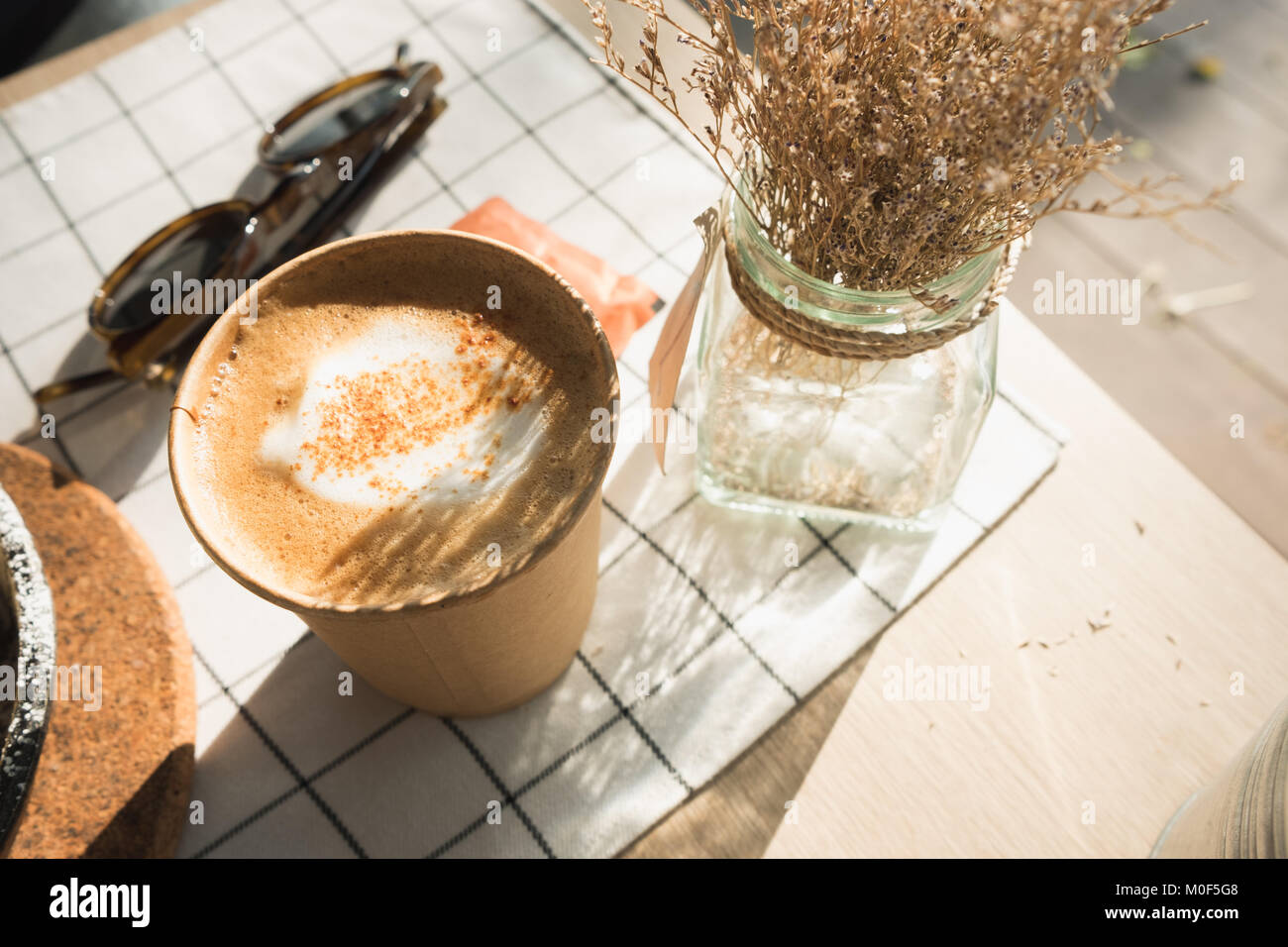 cappuccino in take away coffee cup on table cloth with dry flower,sunglasses on wood table with sunlight at window in evening.food and drink lifestyle Stock Photo
