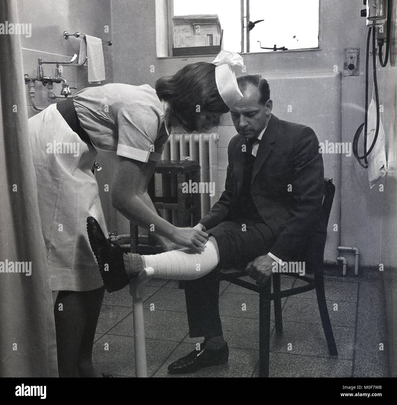 1960s, historical, female nurse putting a dressing or bandage on a gentleman's leg in a GP's surgery, England, UK. Stock Photo