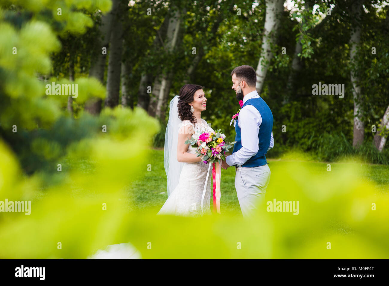Groom and bride smile to each other in the forest with pink flower bouquet and beautiful wedding clothes Stock Photo
