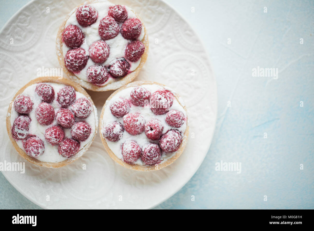 Delicious raspberry tartlets on a white vintage plate. Sweet treat on a light blue background. Flat lay and copy space. Top view. Stock Photo