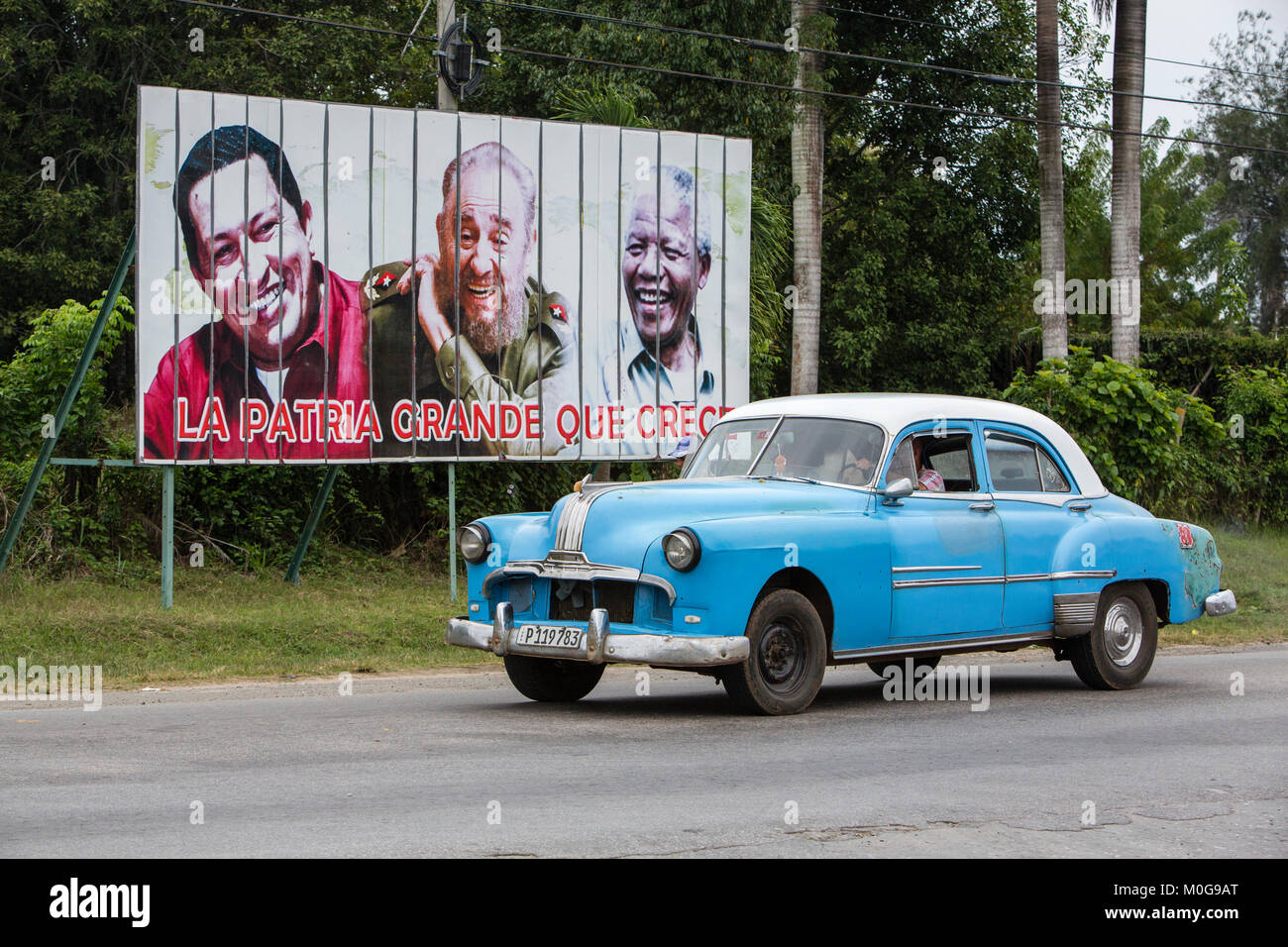Classic American car driving past billboard of Hugo Chavez, Fidel Castro and Nelson Mandella in Cienfuegos, Cuba Stock Photo