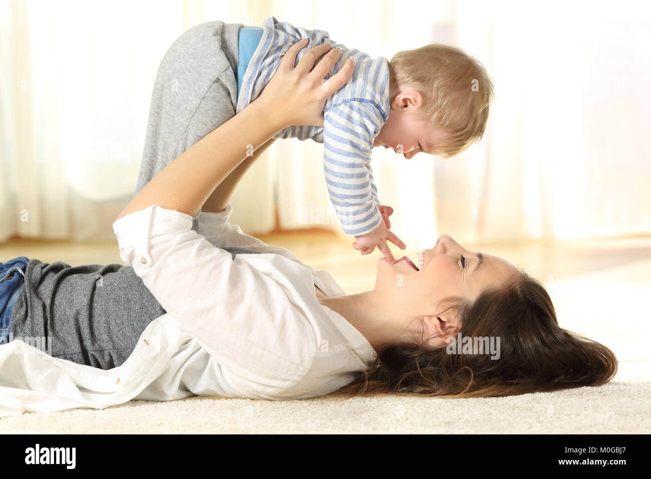 Side view portrait of a happy mother lying on the floor raising her baby son at home Stock Photo
