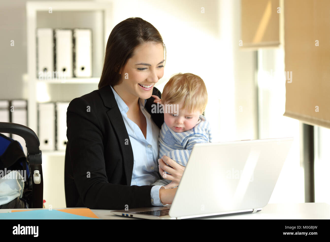 Happy mother working on line with a laptop holding her baby son at office Stock Photo
