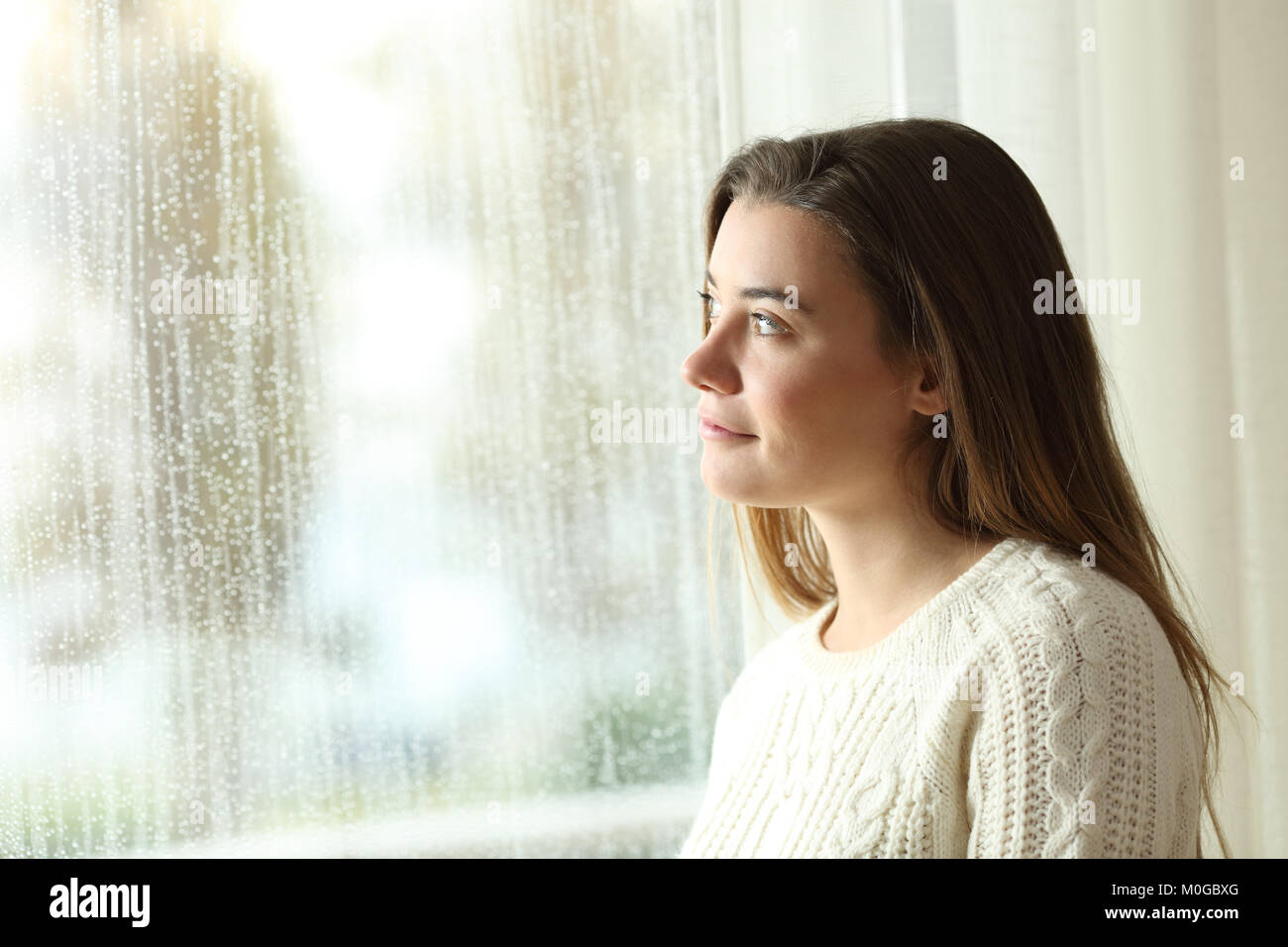 Pensive teenager looking through a window in a rainy day at home Stock Photo