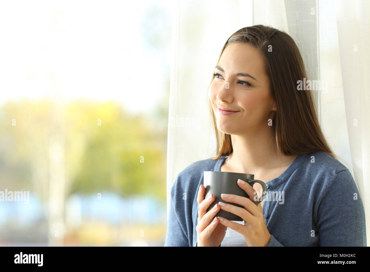Portrait of a satisfied woman thinking looking at side holding a coffee mug beside a window at home Stock Photo