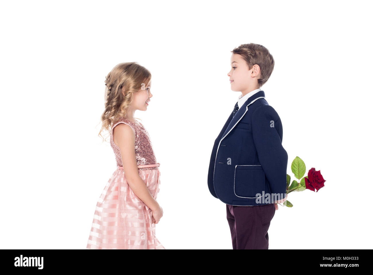 side view of fashionable boy in suit holding rose flower and looking at adorable little girl in pink dress isolated on white  Stock Photo