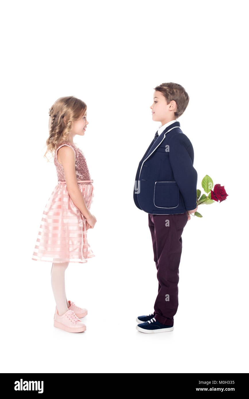 side view of stylish boy in suit holding rose flower and looking at beautiful little girl in pink dress isolated on white  Stock Photo