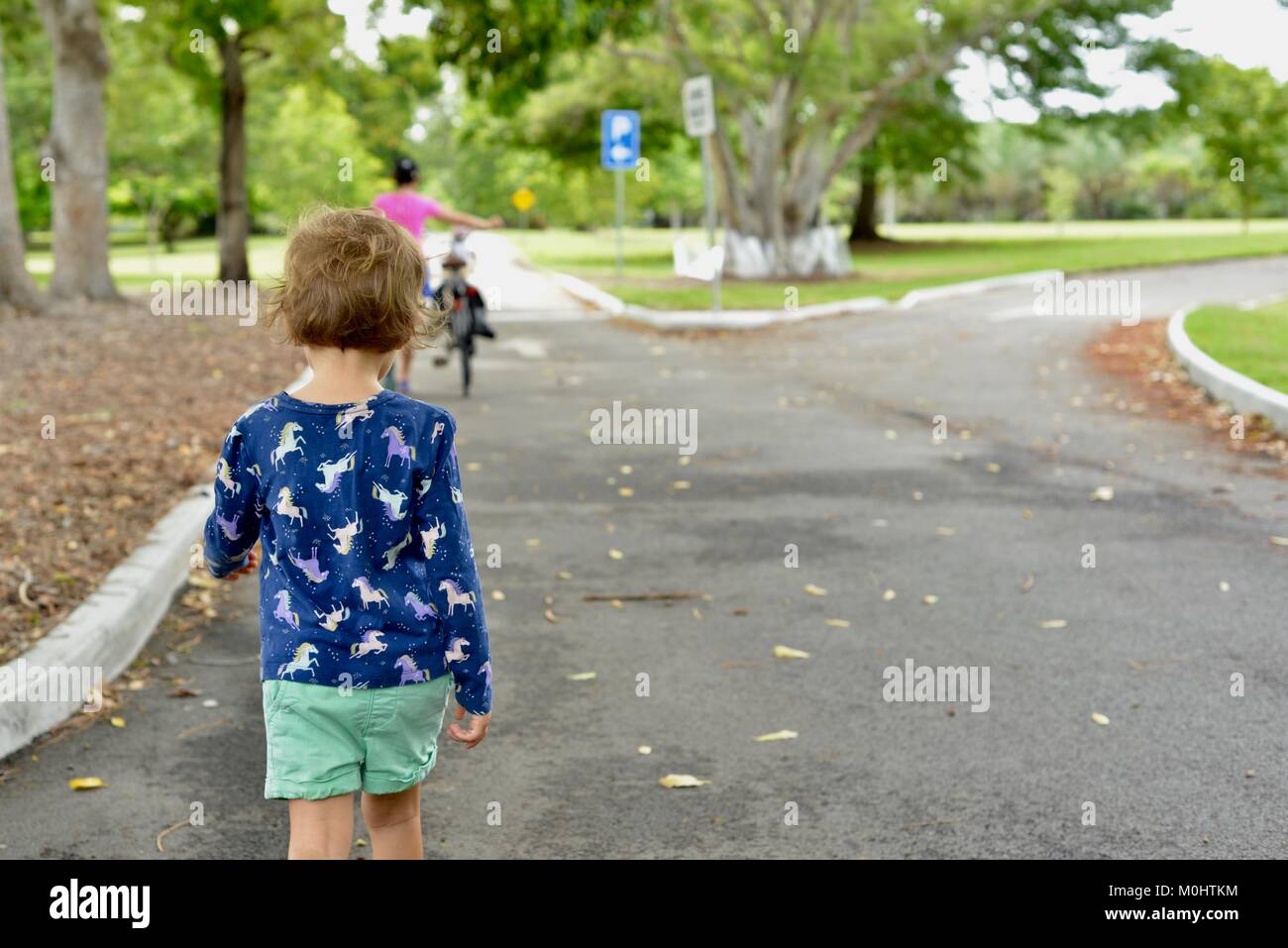 Young child walks on a road with women and bike in distance, Anderson Park Botanic Gardens, Townsville, Queensland, Australia Stock Photo