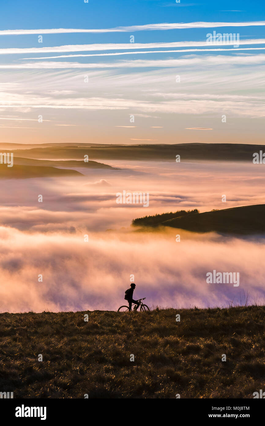 Sun setting over a fantastic Peak District mountain landscape. A biker makes its way through the hills and the fog. A calm yet exhilarating scene. Stock Photo