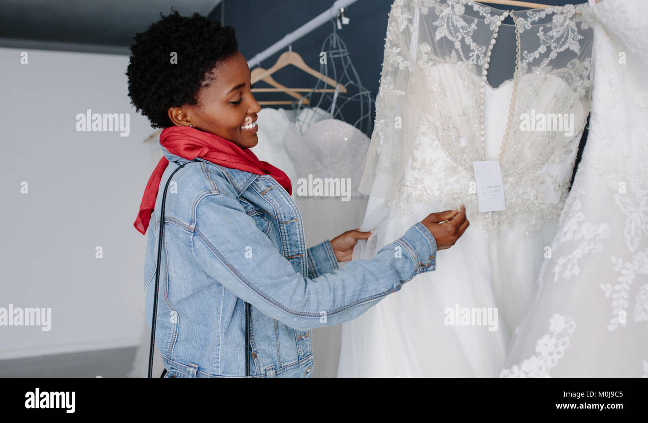 Smiling african woman shopping for wedding outfit in bridal boutique. Bride choosing white gown at shop of wedding fashion. Stock Photo