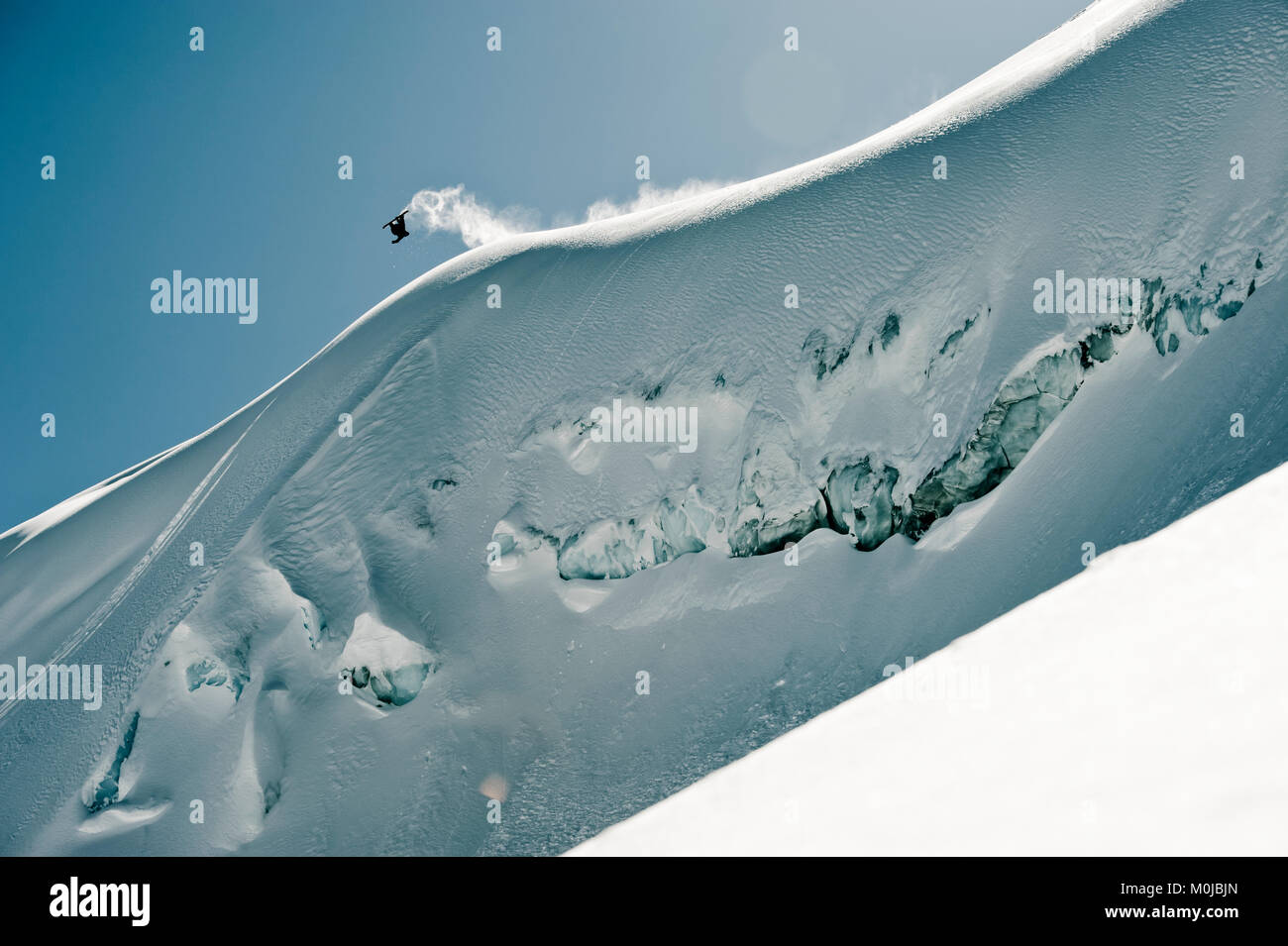 A professional, freeriding snowboarder flips mid-air on a snowy slope against a blue sky; British Columbia, Canada Stock Photo