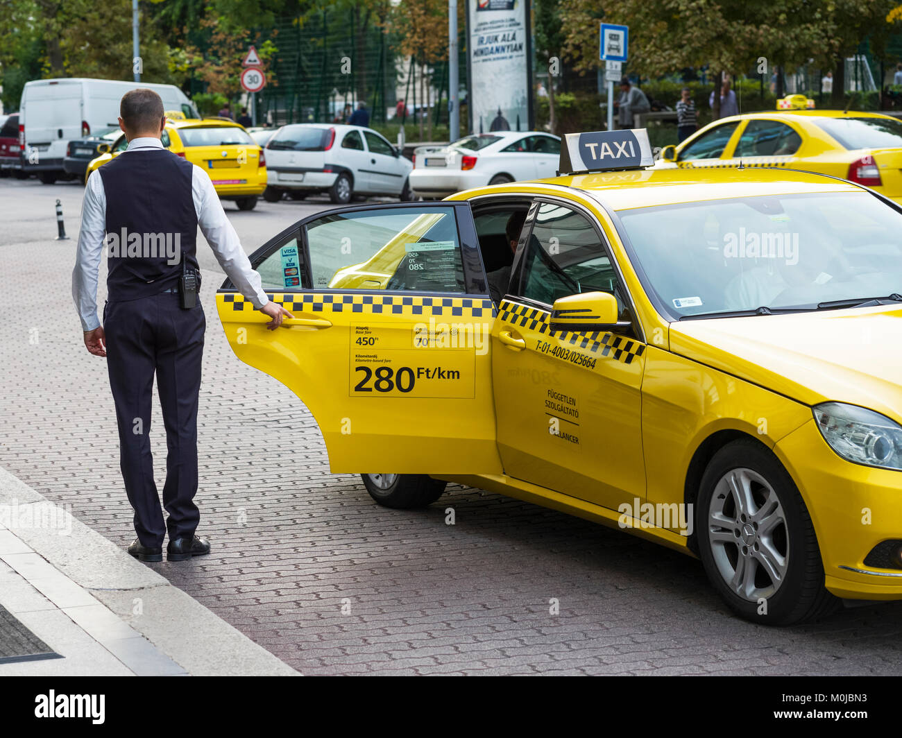 A man stands holding open the door of a yellow taxi waiting for the passenger to exit; Budapest, Budapest, Hungary Stock Photo