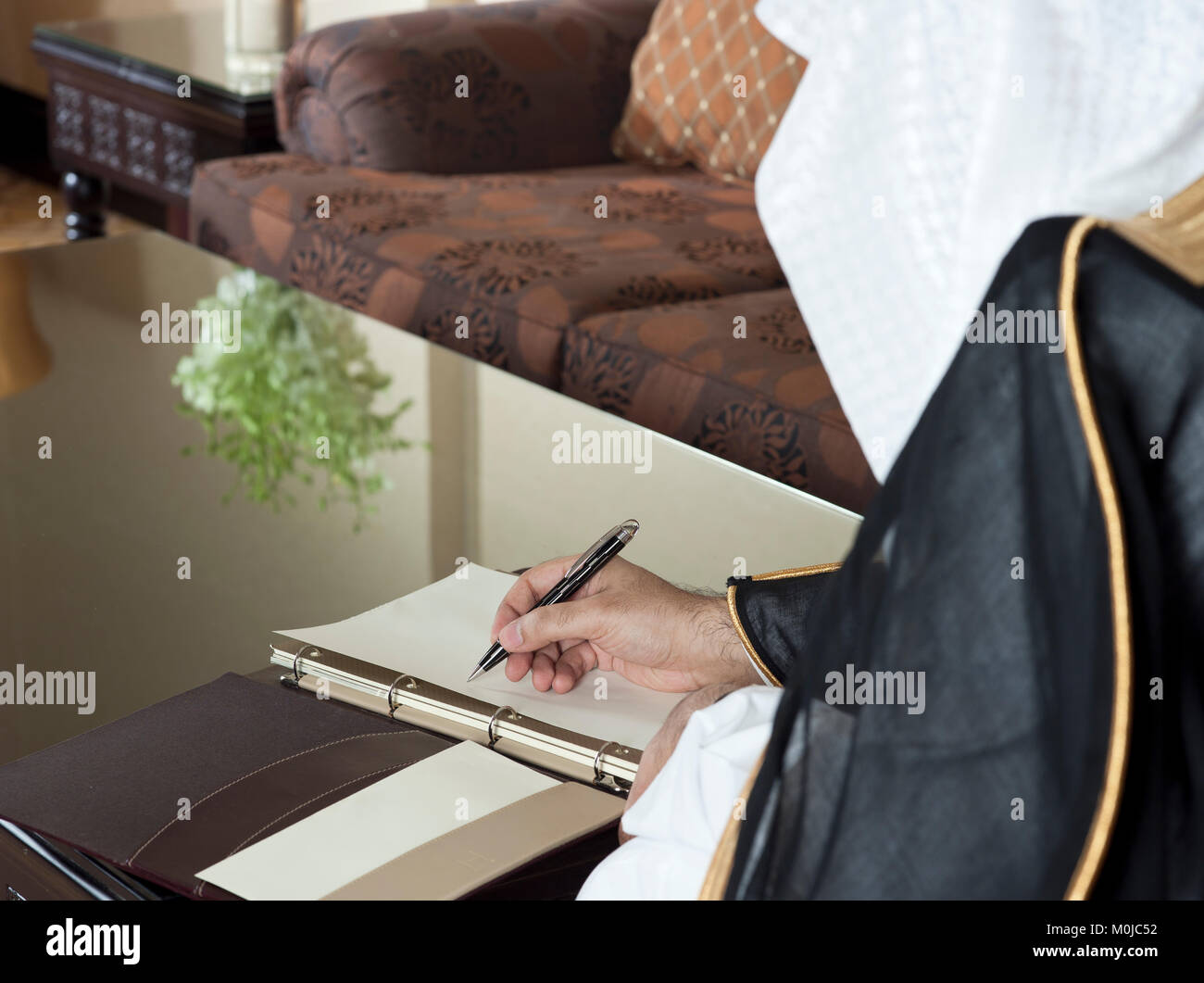 Saudi Arabian Man Hand Writing on A Notebook in a Luxury Home Environment, wearing Saudi Thob, Ghutra and Black Bisht Stock Photo