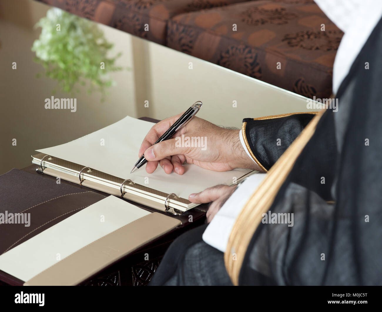 Saudi Arabian Man Hand Writing on A Notebook in a Luxury Home Environment, wearing Saudi Thob, Ghutra and Black Bisht Stock Photo
