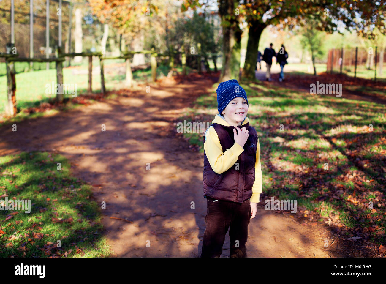 A smiling small boy in warm clothing is ready to have fun in a golden autumn park in United Kingdom. Autumn light, freedom, happiness, childhood. Stock Photo