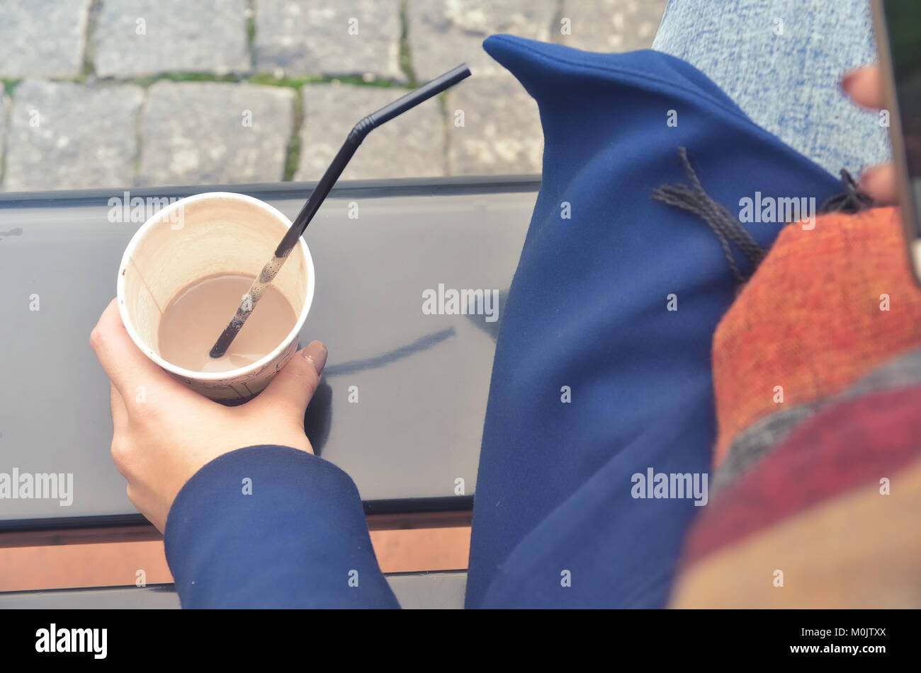 A woman's hand holds a paper cup of coffee. From the cup, stick out the straw. The cup stands on the bench. You can see a paving stone Stock Photo