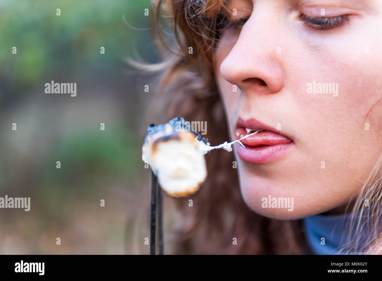 One young woman eating roasted caramelized marshmallow skewer closeup macro portrait showing teeth, biting, mouth, face, tongue Stock Photo