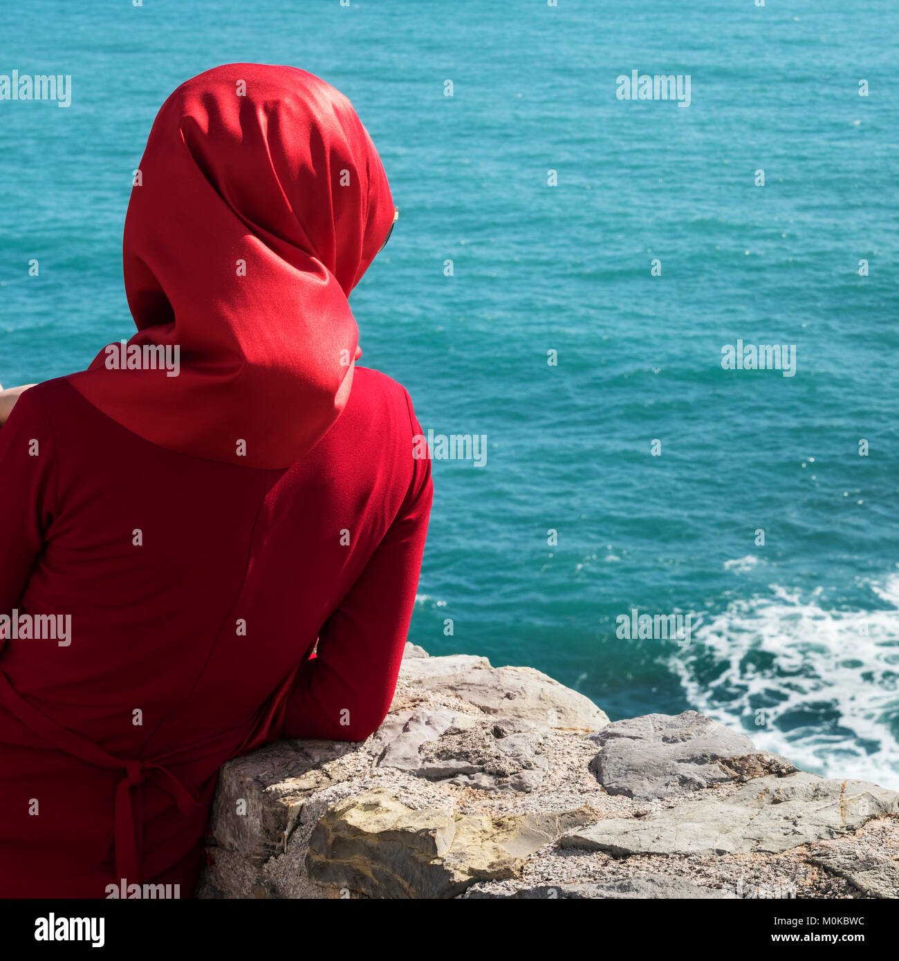 A woman wearing a red headscarf and red dress stands at a stone wall looking out at the blue water; Budva, Budva Municipality, Montenegro Stock Photo