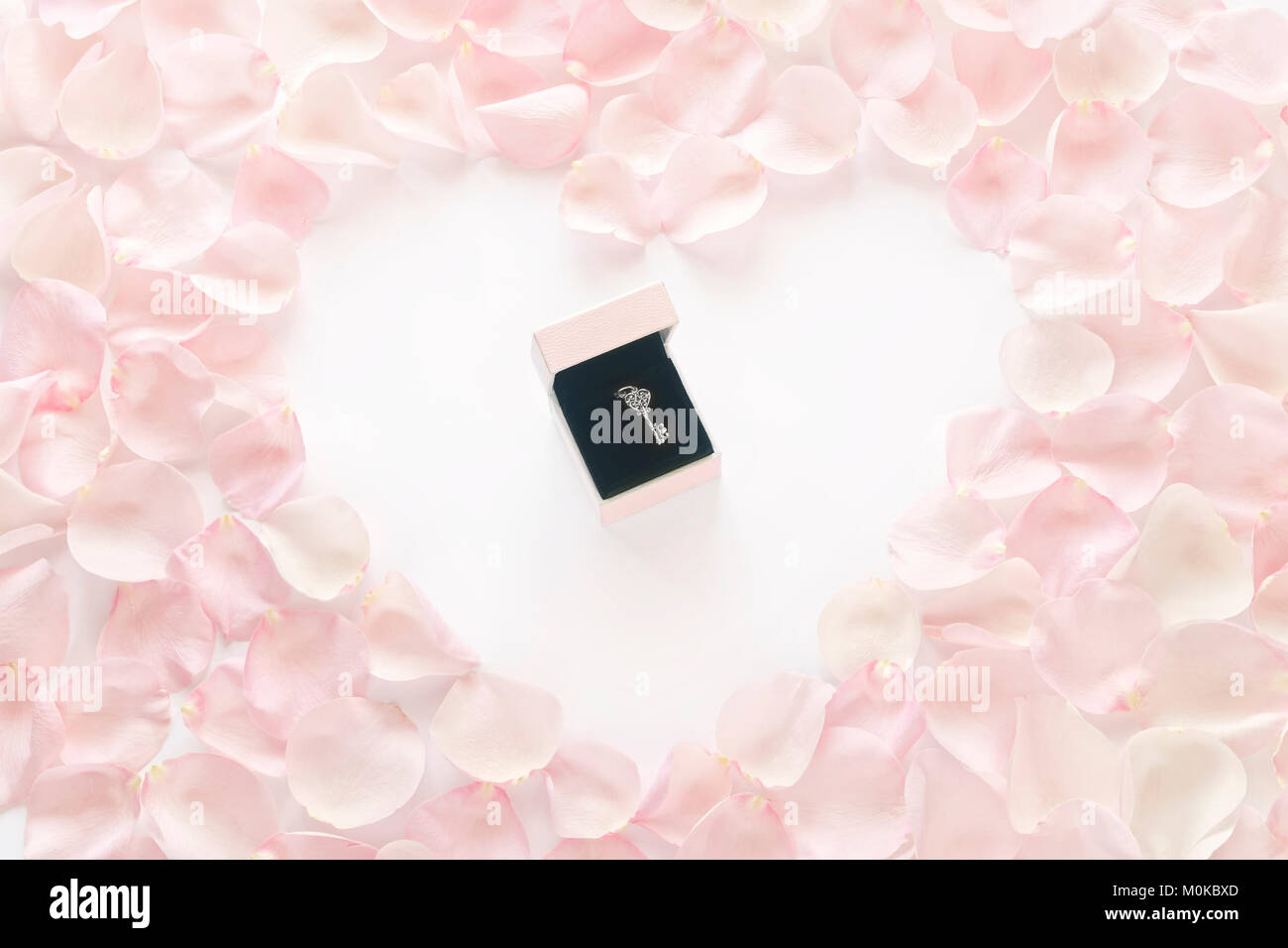 silver key pendant in open white and pink gift box in the middle of heart shaped rose petals on white background Stock Photo