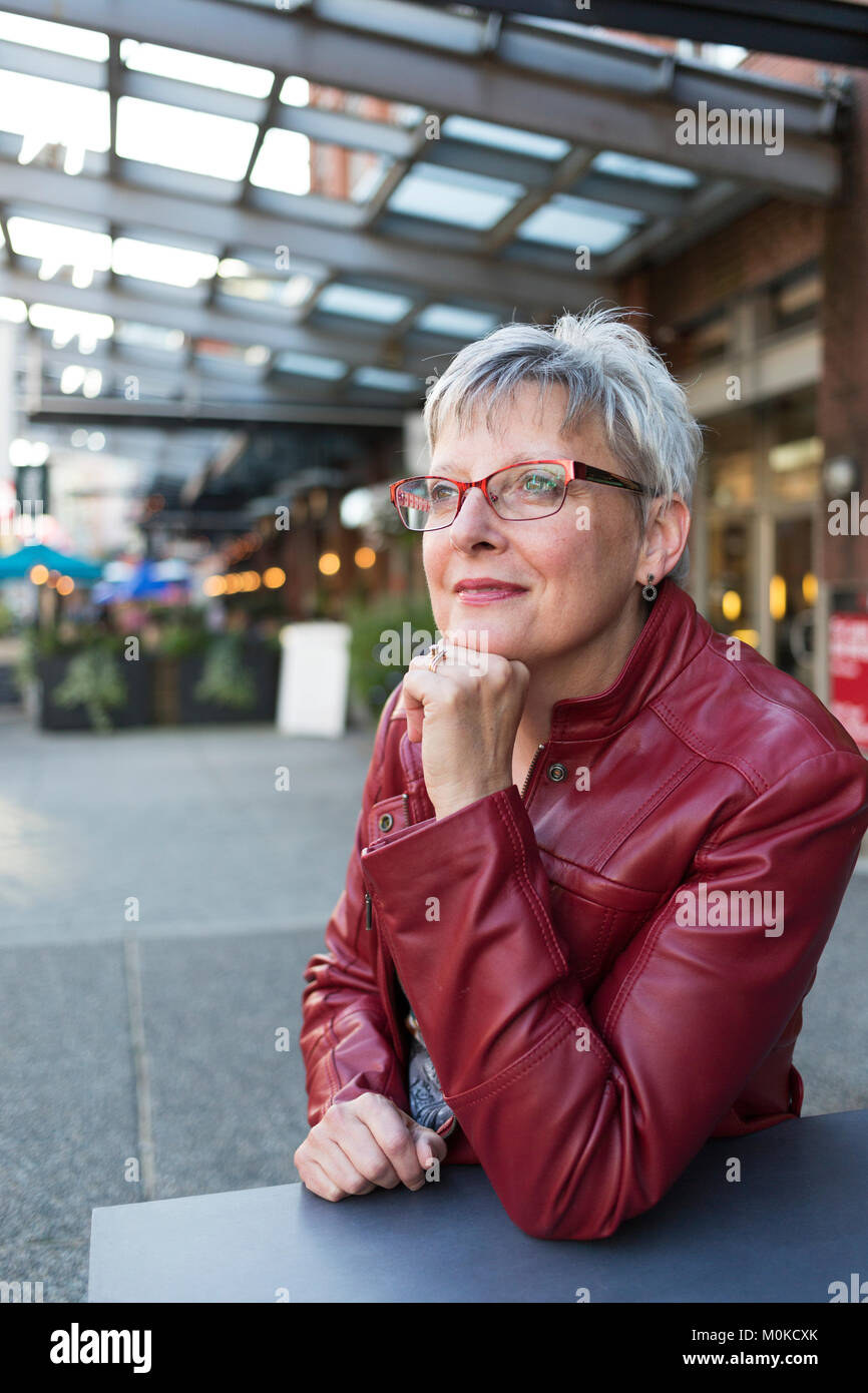 Portrait of a mature woman in a red leather jacket sitting at a table outside a shop with her chin resting in her hand Stock Photo
