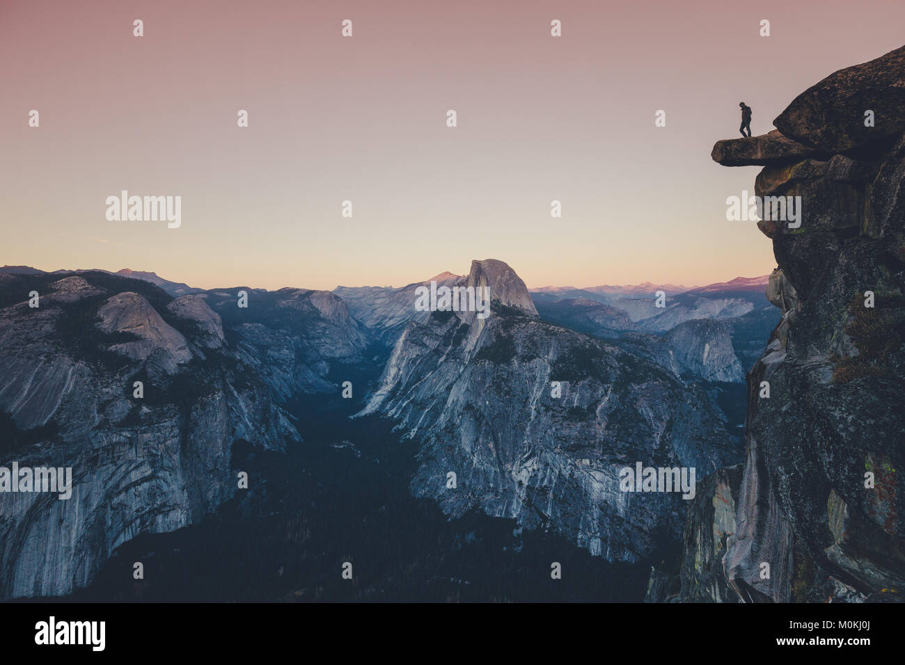 A fearless hiker is standing on an overhanging rock looking towards famous Half Dome in Yosemite National Park in twilight at sunset, California, USA Stock Photo