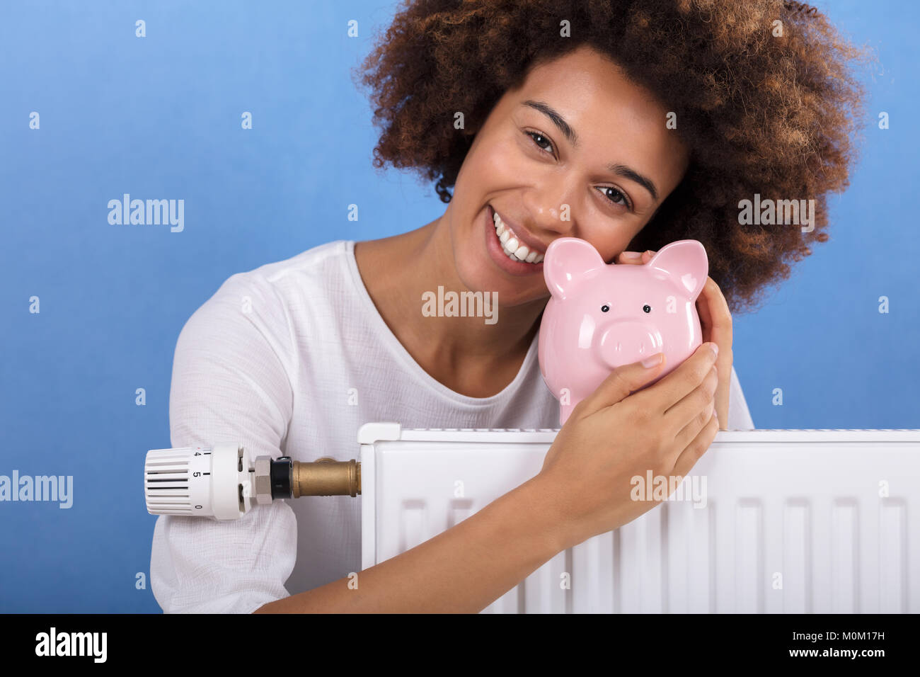 Happy Young Woman Behind The Heating Radiator Holding Pink Piggy Bank Stock Photo
