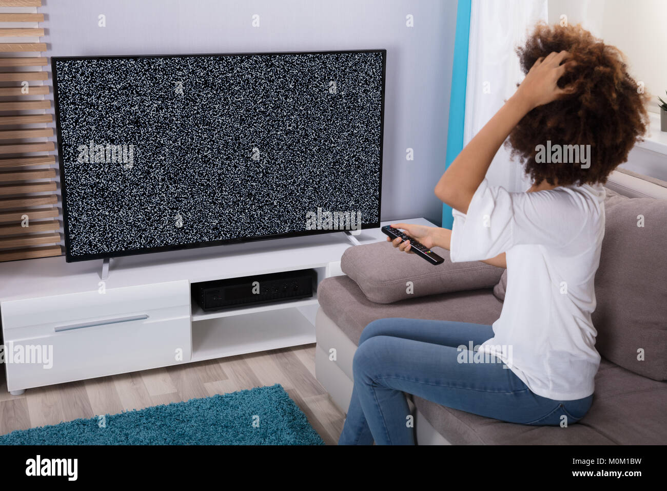 Young Woman Sitting On Sofa Near Television Showing No Signal Stock Photo