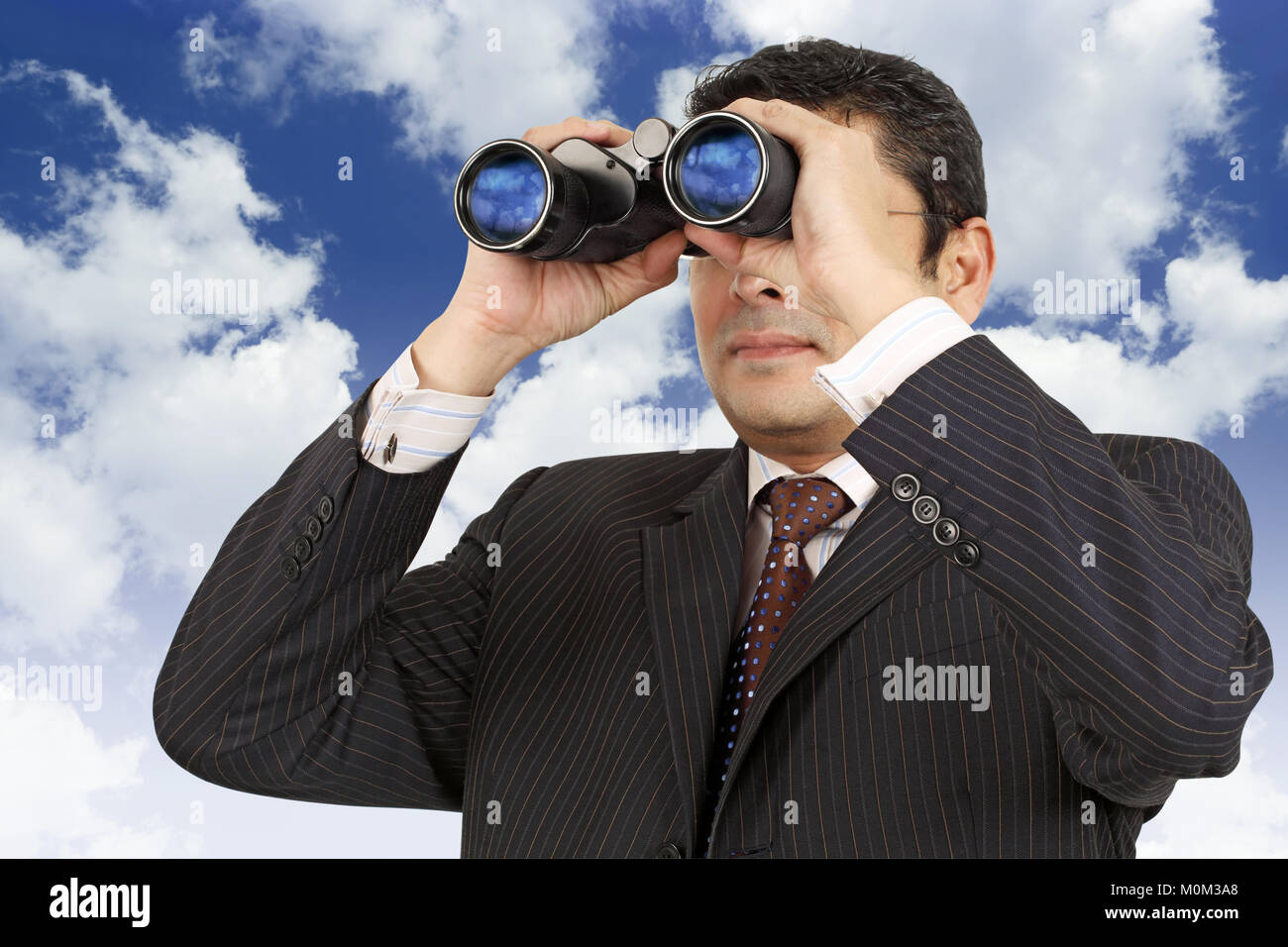An Indian businessman in his late thirties looking through binoculars with a cloudy blue sky in the background. Stock Photo