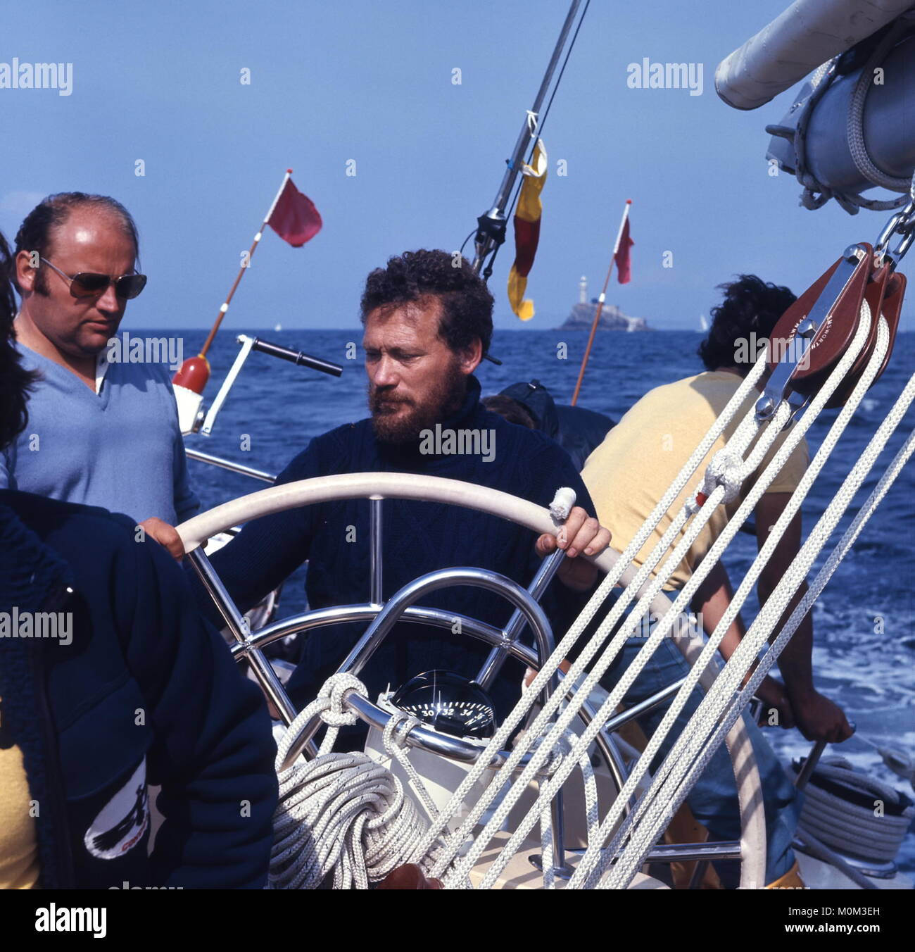 AJAX NEWS PHOTOS. 1977. IRISH SEA. - FASTNET RACE SKIPPER - ROBIN KNOX-JOHNSTON (GB) AT THE WHEEL OF THE BOWMAN 77 YACHT CONDOR DURING THE FASTNET RACE, THE ICONIC LIGHTHOUSE ASTERN. YACHT OWNER BOB BELL ON LEFT.   PHOTO:JONATHAN EASTLAND/AJAX REF:703734 14 Stock Photo