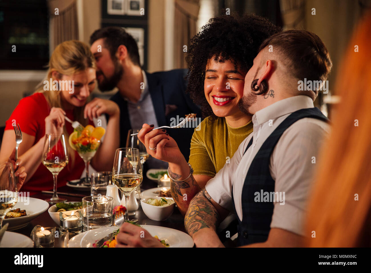 Man is kissing his partner on the cheek while they are dining at a restaurant with other friends. Another couple can be seen out of focus in the backg Stock Photo