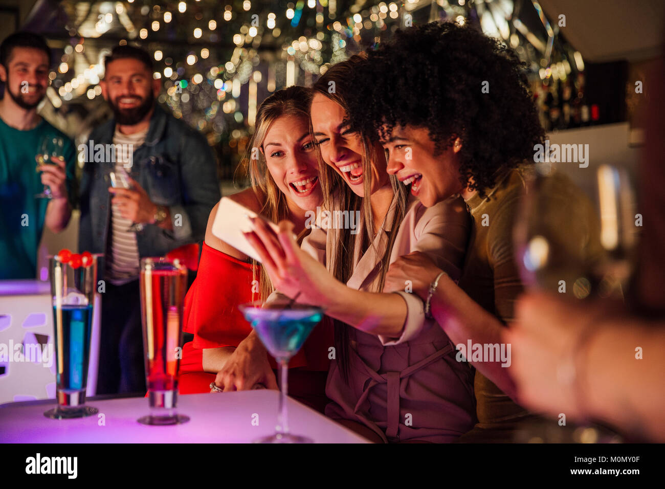 Three women are taking selfies together on a smartphone while enjoying drinks in a night club. Stock Photo