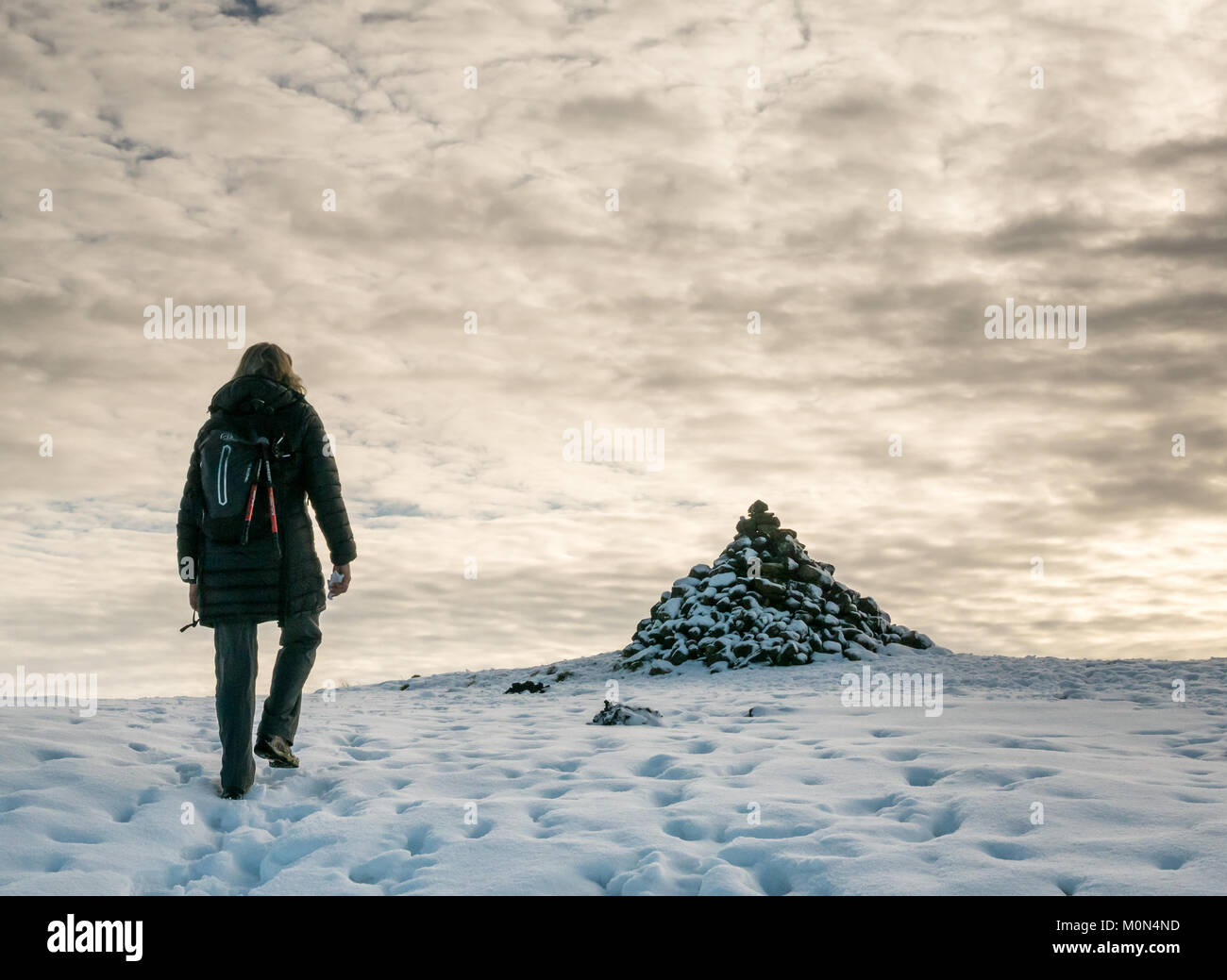 Woman walking in snow uphill on footpath between Langbank and Kilmacolm towards snow covered cairn, Strathclyde, Scotland, UK, with mottled cloudy sky Stock Photo