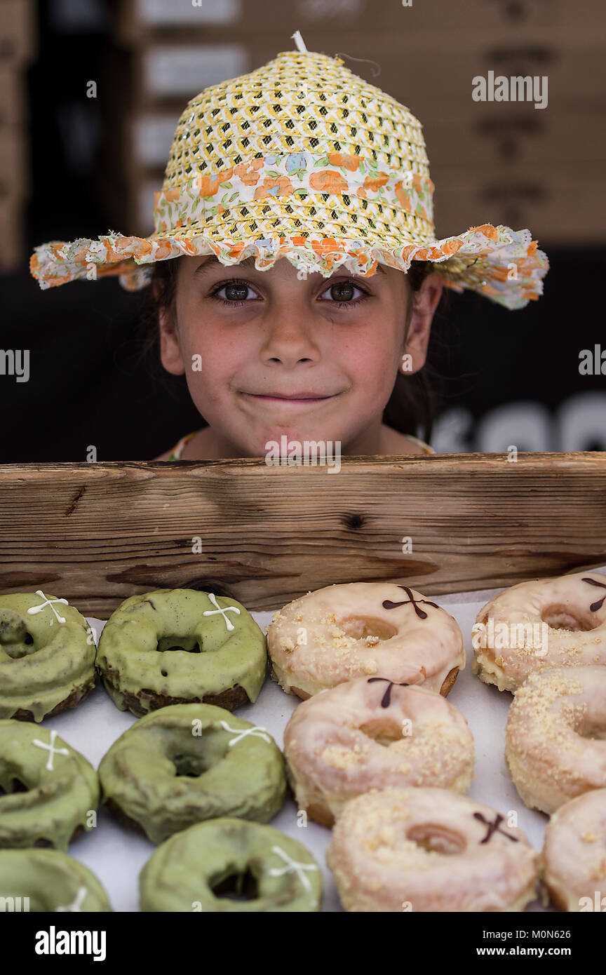 Young girl with a quirky smile watching green artisan donuts quite closely and wearing a straw hat at a street festival in Marlybone London UK Stock Photo