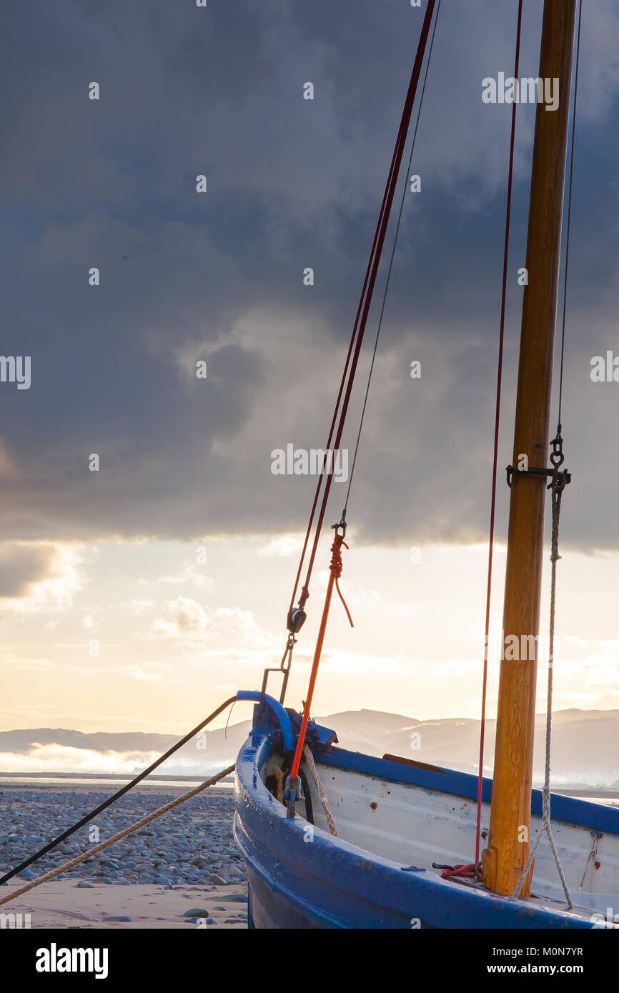 Portrait close up of blue fishing boat anchored on Ynyslas Beach. Sea & sunlit mountains in background. Dramatic scene: dark foreboding storm clouds. Stock Photo