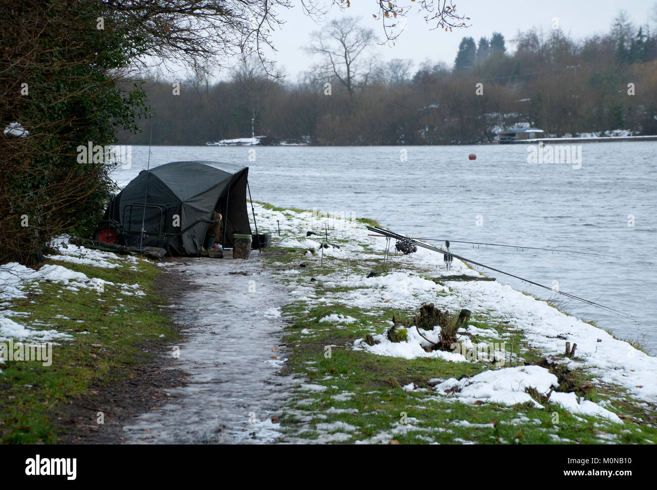 Fishing on a cold day with snow and ice on the ground. Angler sitting in a fishing shelter or tent while fishing a lake in England. Stock Photo