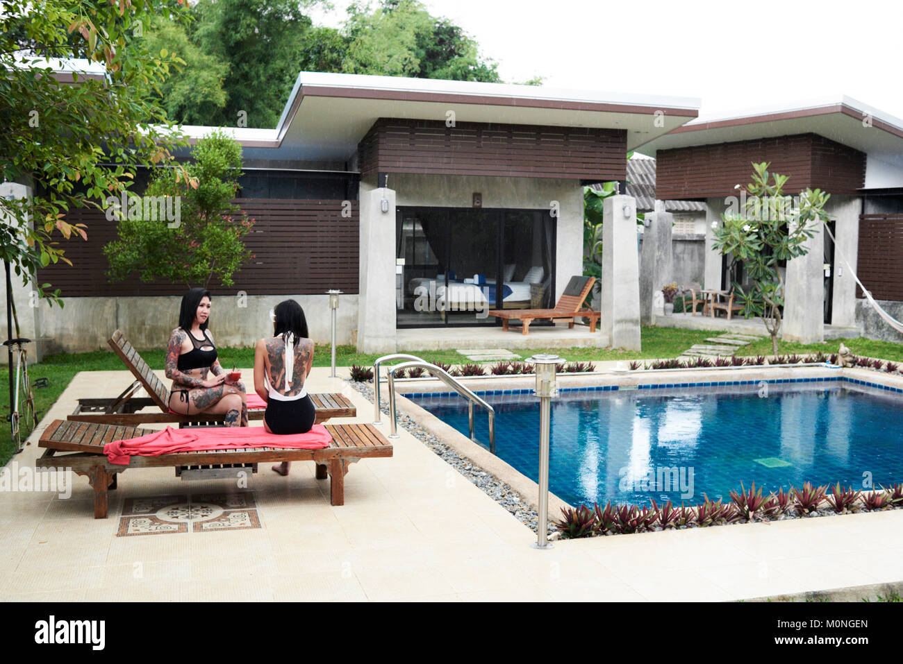 Wide view of tattooed asian friends having fun and drinking next to the swmming pool while raining. Stock Photo